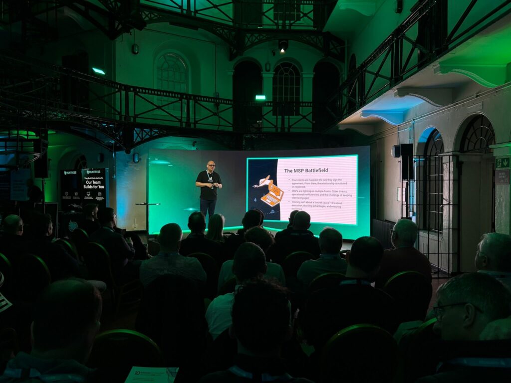 Speaker presenting at the Huntress Roadshow. Green and blue lighting fills the room.